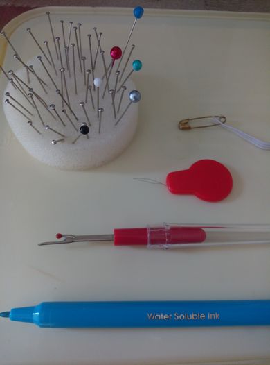 Left to right and top to bottom: Cushion of pins, safety pin and elastic, needle threading tool, pick tool, water-soluble marker