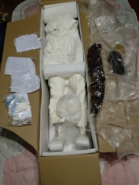 Filler material and goodies put to the side. You see the 3 clothes items, 2 wigs, goodiebag. Inside one can see the syrofoam blocks that should help avoid moving, especially the legs. You see the doll is wrapped in a blanket.