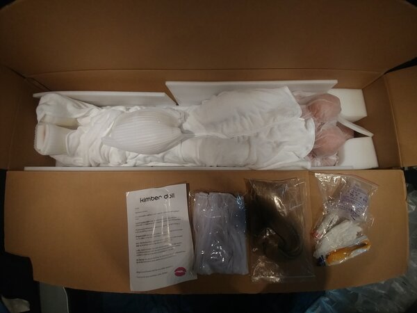 Air blister packaging removed (head is placed at her knees) and included accessories laid out. (Greeting/care letter, lingerie, blonde wig, and accessories kit [warmer rod, comb, linen gloves, irrigator])